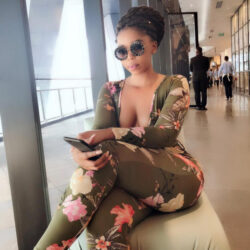 Woman in a green floral jumpsuit and round sunglasses sits by a window in a modern hallway, scrolling on her phone.