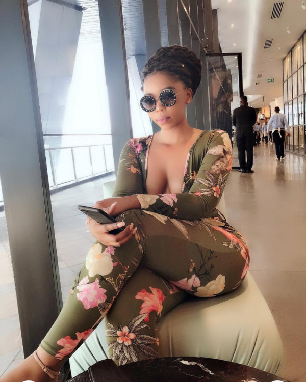 Woman in a green floral jumpsuit and round sunglasses sits by a window in a modern hallway, scrolling on her phone.